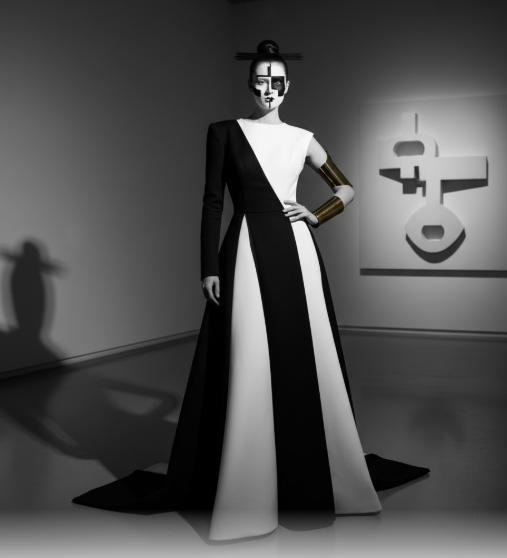 A striking, full-length, black and white image of a figure dressed as a Modernist Minimalist Halloween costume. The costume features a dramatically flared gown with a high contrast, geometric black and white panel design, creating a sculptural silhouette that suggests architecture in motion. The figure's face is painted with a bold, graphic, abstract design in black and white, and her hair is styled in a severe, minimalist bun. A single, prominent gold cuff adorns one arm. The background is a minimalist gallery space with a large, white, abstract sculptural artwork on the wall, reinforcing the architectural and artistic theme. The overall aesthetic is monochrome, bold, and avant-garde.