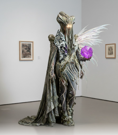 A refined, visually attractive image of a figure in a Quiet Couture Creature costume. The garment is a sculptural, flowing dress in layered, sheer fabrics of charcoal and dusty rose, creating an experimental yet composed silhouette. One arm features an elegant arrangement of sheer, winged appendages or fins. The face is visible but subtly masked with gold leaf detailing, and the figure holds a translucent, glowing white sphere. The look blends creature features with high-fashion elegance, prioritizing sophisticated allure.