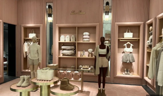 Interior of a Loro Piana luxury boutique installation, possibly at Bergdorf Goodman, featuring a monochromatic palette of blush pink and beige. Mannequins display cashmere loungewear and accessories on plush, gold-accented displays.