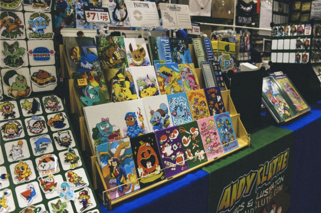 Artist alley booth displaying colorful character stickers, art prints, and bookmarks on tiered racks. 