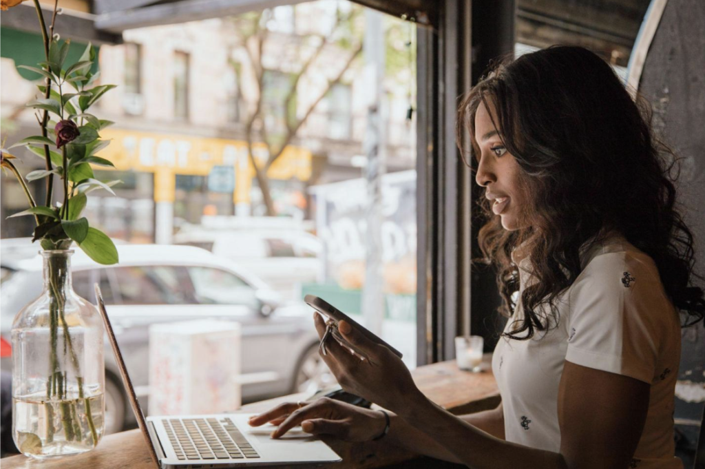 Woman using a laptop while holding a smartphone at a café table. 