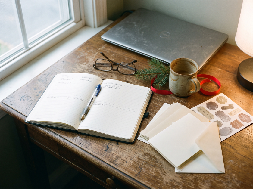 An open planner, blank holiday cards and a mug of coffee on a wooden desk by a window